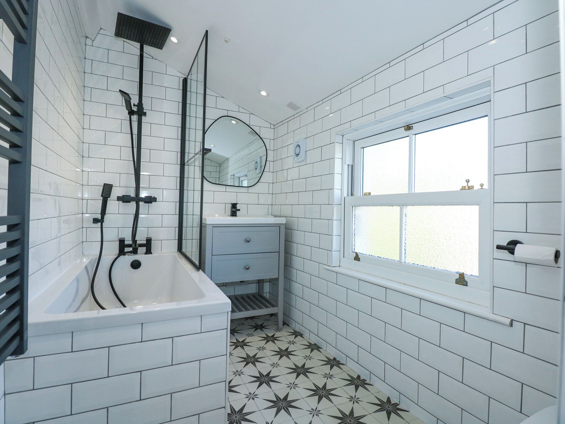 Un baño moderno con azulejos blancos tipo metro, una bañera independiente, un lavabo con un espejo encima y una ventana con vidrio esmerilado.
