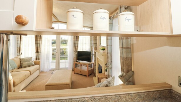 Ein modernes Wohnzimmer mit einer Couch, einem Couchtisch und einem Fernseher. Auf einem Holzregal befinden sich drei weiße Aufbewahrungsbehälter mit den Aufschriften 'Tee', 'Kaffee' und 'Zucker'.