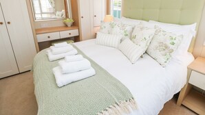 Ein ordentlich eingerichtetes Schlafzimmer mit Bett, Kissen, Nachttisch, Spiegel, Fenster mit Jalousien und einem an der Wand befestigten Regal.