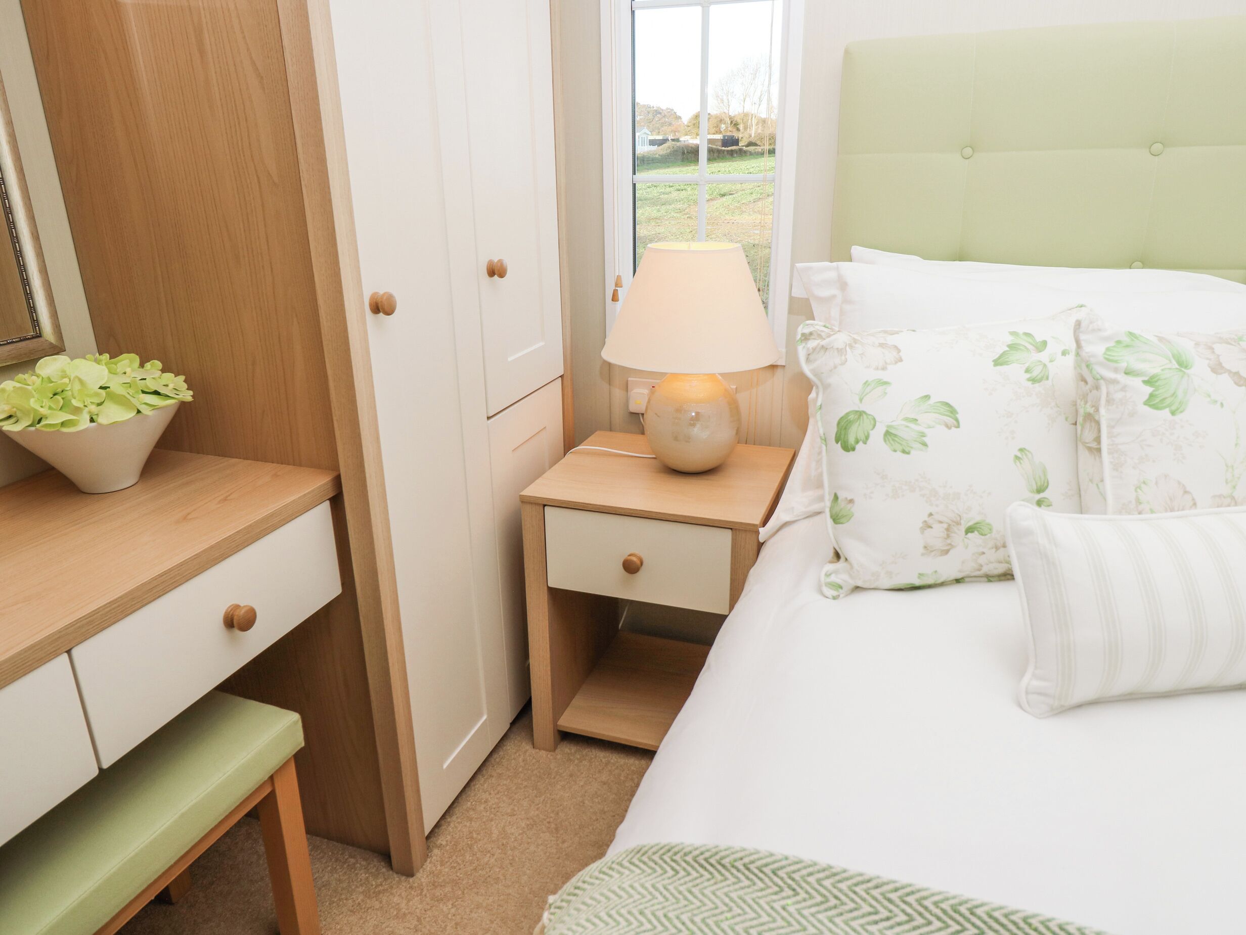 Un dormitorio ordenado con cama, mesita de noche, lámpara y un pequeño banco.