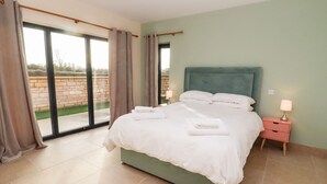 A bedroom with a large bed, a nightstand with a lamp, and a view of a garden through a sliding glass door. - Bears Court Barn 1 (Cheltenham)