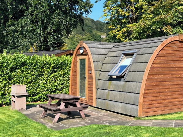 A small, wooden cabin with a sloped roof and a skylight. It is surrounded by lush greenery and has a picnic table in front. - Dovestone Pod 24 (Oldham)