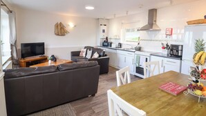 A modern living room with a sofa, TV, and dining table with chairs. The kitchen area includes a stove, sink, and refrigerator. - Castle View Lodge (Ilfracombe)