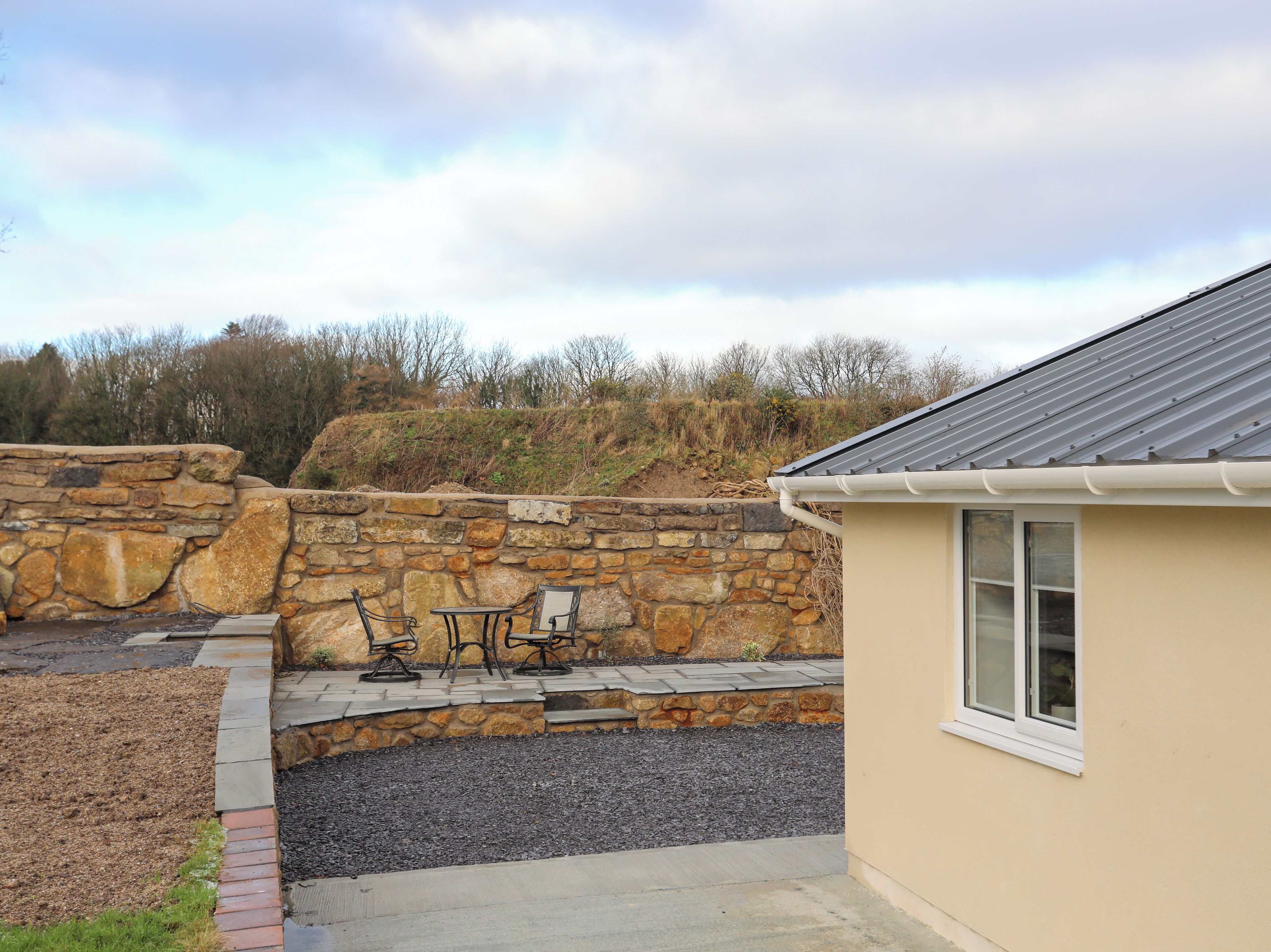 A stone wall, a small patio with a table and chairs, and a building with a window.