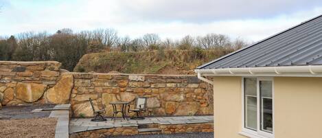 A stone wall, a small patio with a table and chairs, and a building with a window.