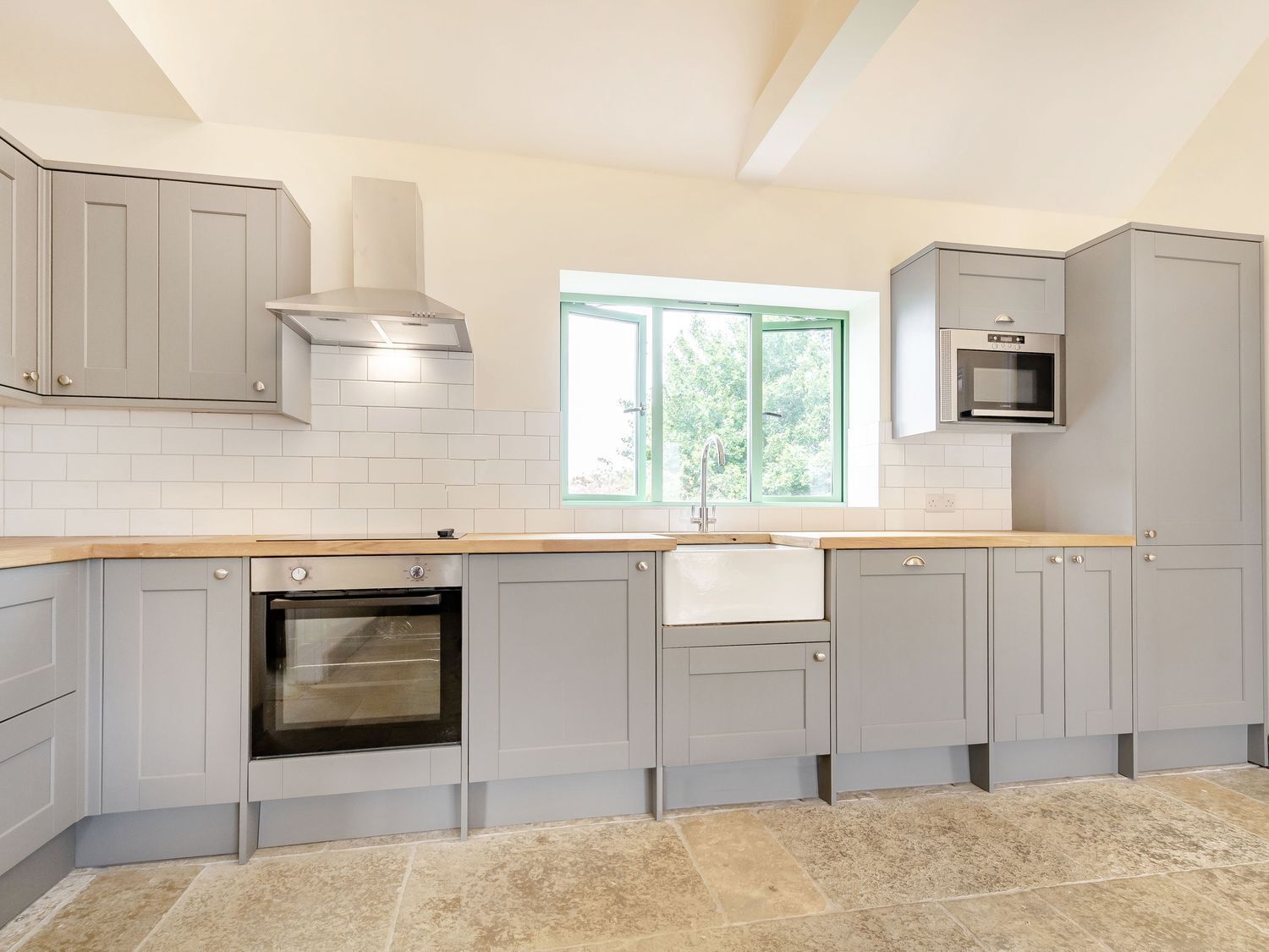 A modern kitchen with grey cabinets, a built-in oven, a stainless steel range hood, a window with a view of greenery, and a microwave.