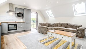 A modern kitchen with grey cabinets, a microwave, and an oven. There is a grey carpet and a wooden coffee table. - Bears Court The Flat (Cheltenham)