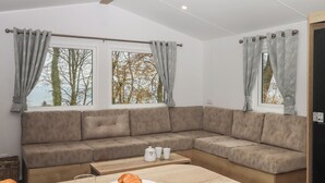 A modern living room with a sectional sofa, two windows with patterned curtains, a wooden coffee table with croissants and a glass of orange juice, and a small side table with a kettle and cups. - 6 Manor Park (Bideford)