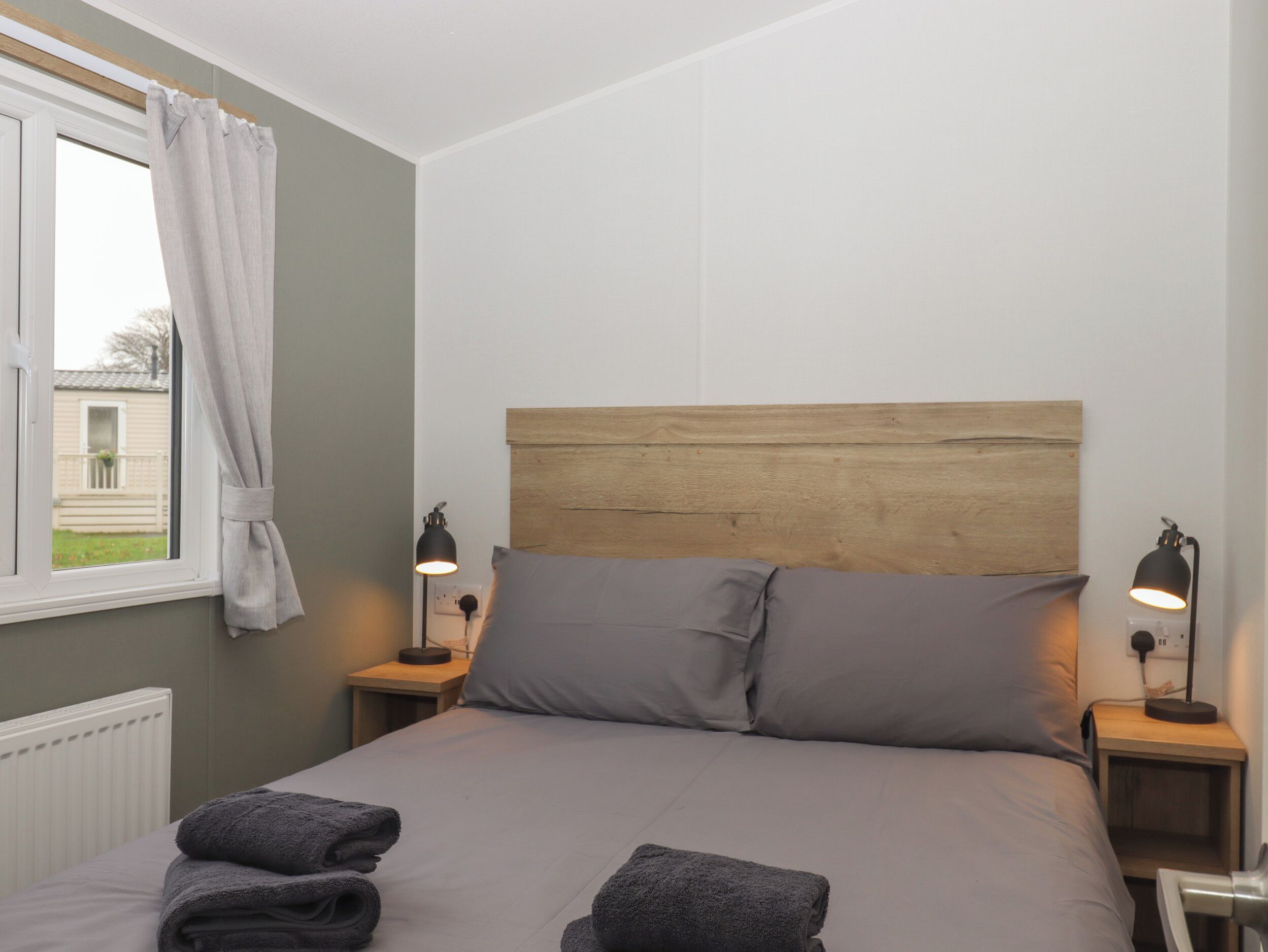 A neatly made bed with grey bedding and two pillows, two bedside lamps, and a window with a curtain.