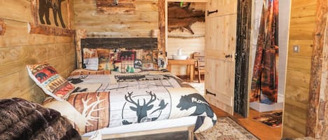 A rustic cabin interior with a bed, pillows with animal prints, a wooden ceiling, and a dining area.