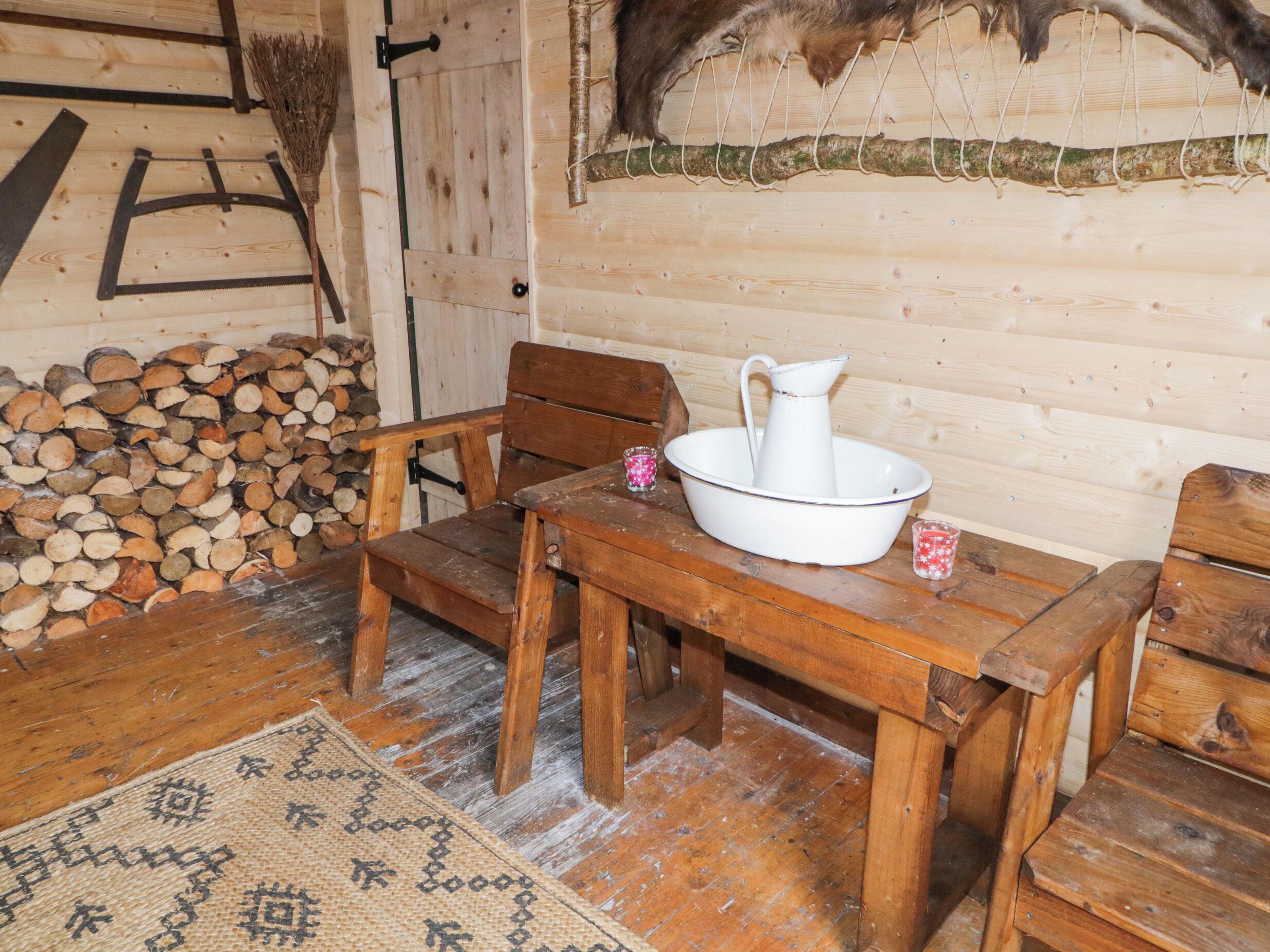 A rustic wooden cabin interior with a wooden table, two wooden chairs, a woven rug, and a pile of firewood against the wall.