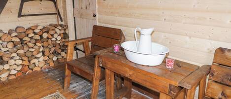 A rustic wooden cabin interior with a wooden table, two wooden chairs, a woven rug, and a pile of firewood against the wall.