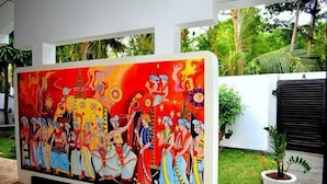 Un mural colorido que muestra figuras tradicionales y un templo, sobre un muro blanco con una planta en maceta cerca.
