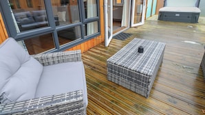 A wooden deck with a sofa and a wicker ottoman, a sliding glass door leading to a room, and a mattress on the deck. - Dovestone Luxury Lodges - Pets Welcome (Oldham)