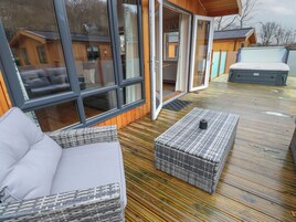A wooden deck with a sofa and a wicker ottoman, a sliding glass door leading to a room, and a mattress on the deck.