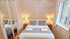 A cozy bedroom with a large bed, two bedside tables with lamps, a window with curtains, and a wooden floor. - Dovestone Secluded Luxury Lodges - Pets Welcome (Oldham)