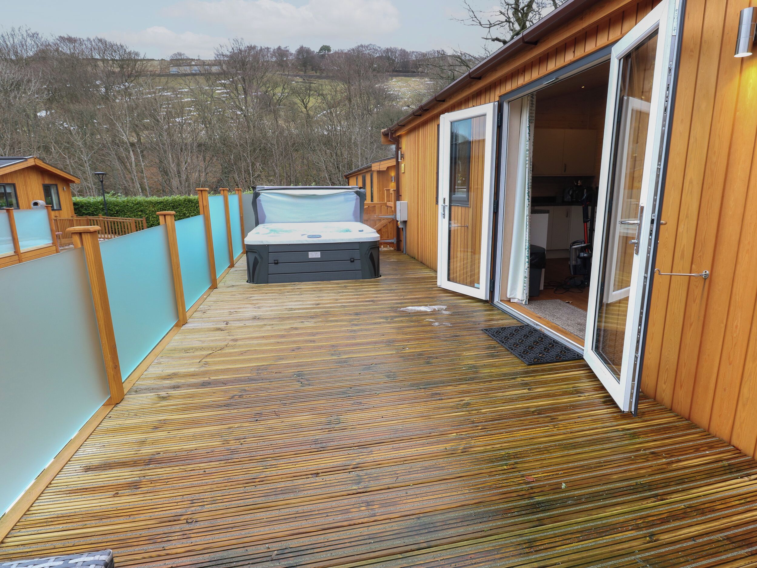 Une terrasse en bois mène à une pièce dotée d’une porte-fenêtre coulissante, d’un jacuzzi et offrant une vue sur un paysage vallonné.