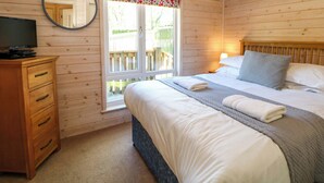 A wooden cabin bedroom with a bed, bedside table, lamp, TV, and a window with a view of a wooden deck. - Dovestone Luxury Lodges - pet Free (Oldham)