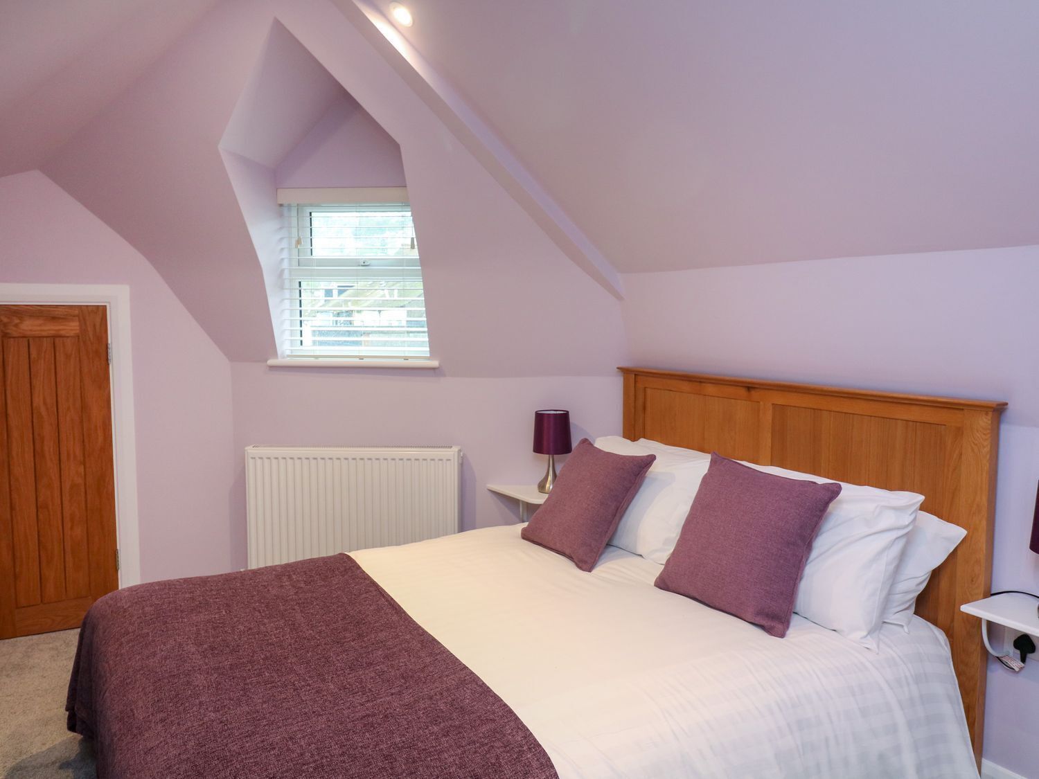 Une chambre avec un plafond en pente, une fenêtre, un radiateur, un lit avec une literie blanche et violette, et une tête de lit en bois.