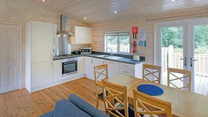 A modern kitchen with white cabinets, a gas stove, and a microwave. There is a dining area with a wooden table and chairs. - Six Berth pet Free Lodges (Oldham)