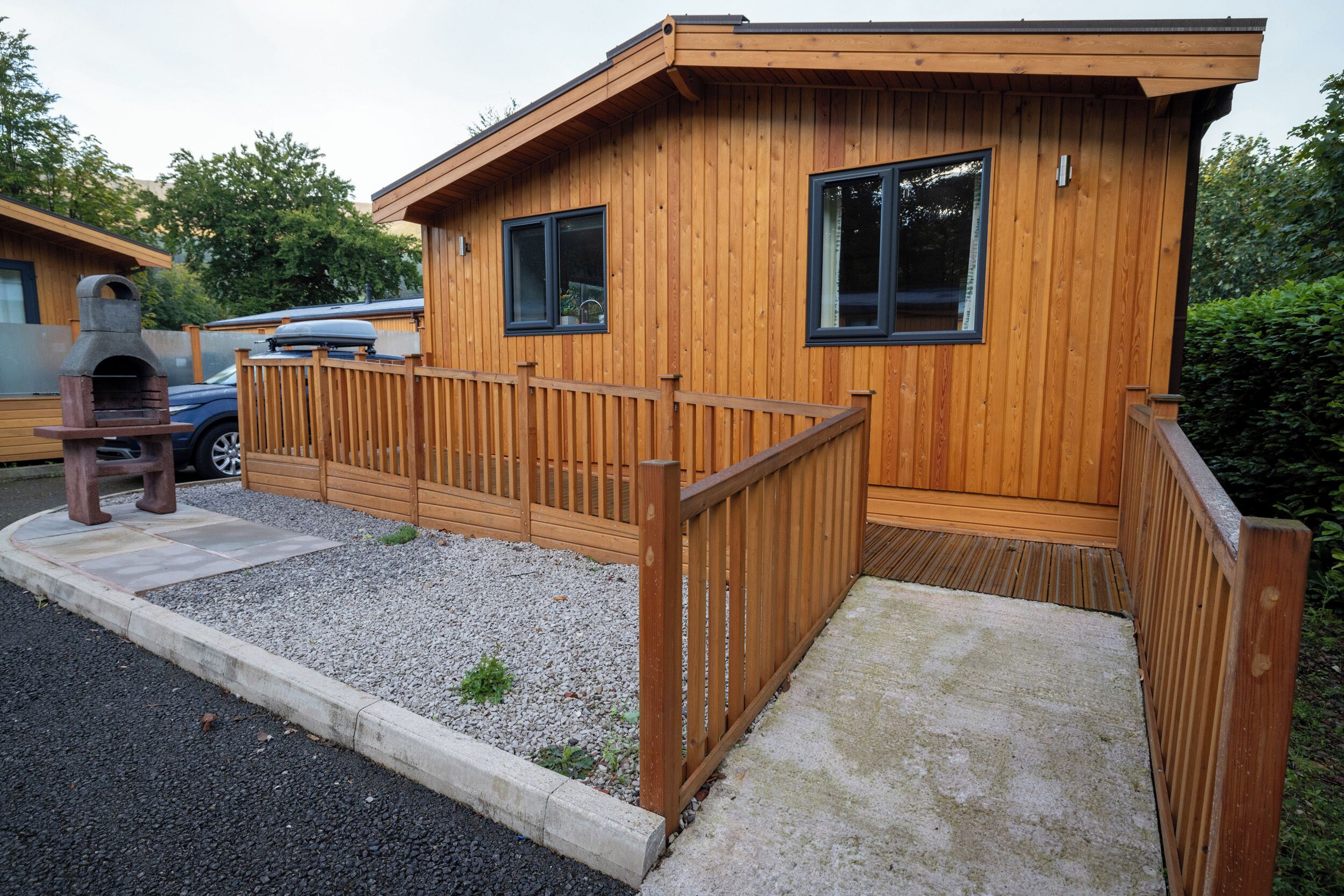 A wooden cabin with a deck, a barbecue area, and a gravel driveway.
