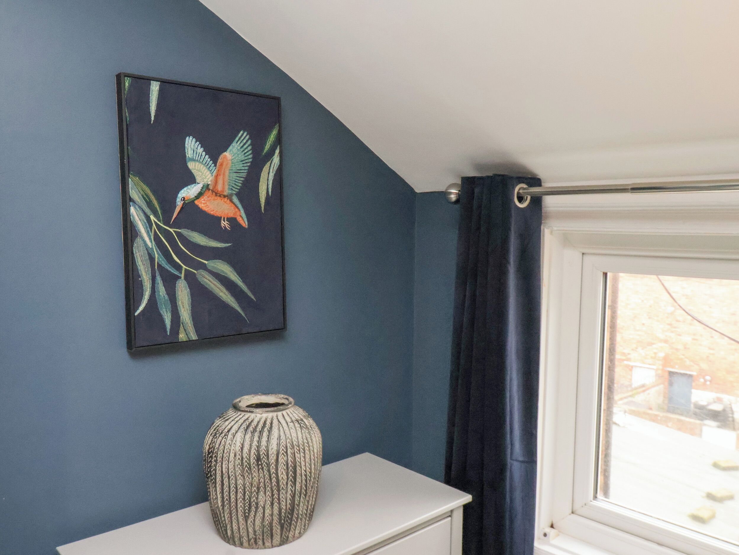 A room with a dark blue wall, a framed painting of a bird, a textured vase, and a window with a curtain.