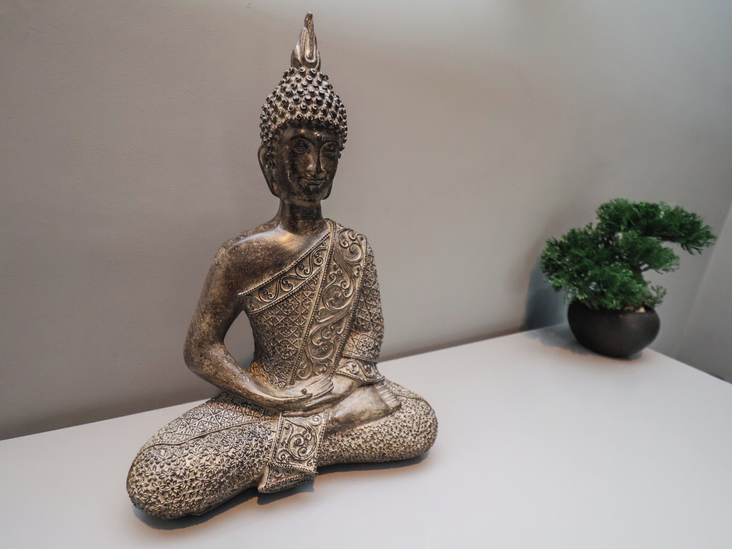 A bronze Buddha statue with intricate patterns, seated on a white surface next to a potted plant.