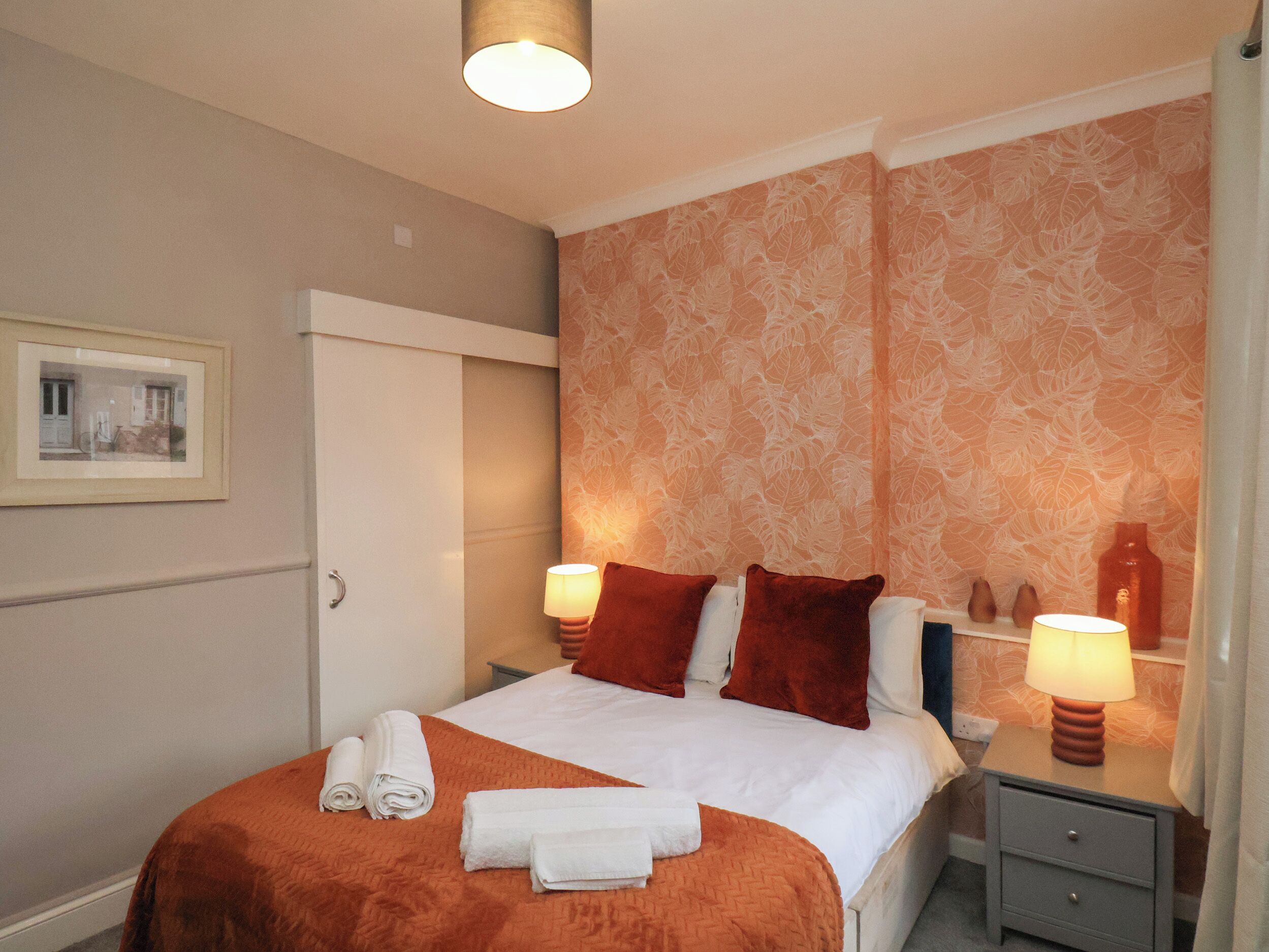 A neatly made bed with white and orange bedding, two bedside lamps, and a small nightstand with a lamp.
