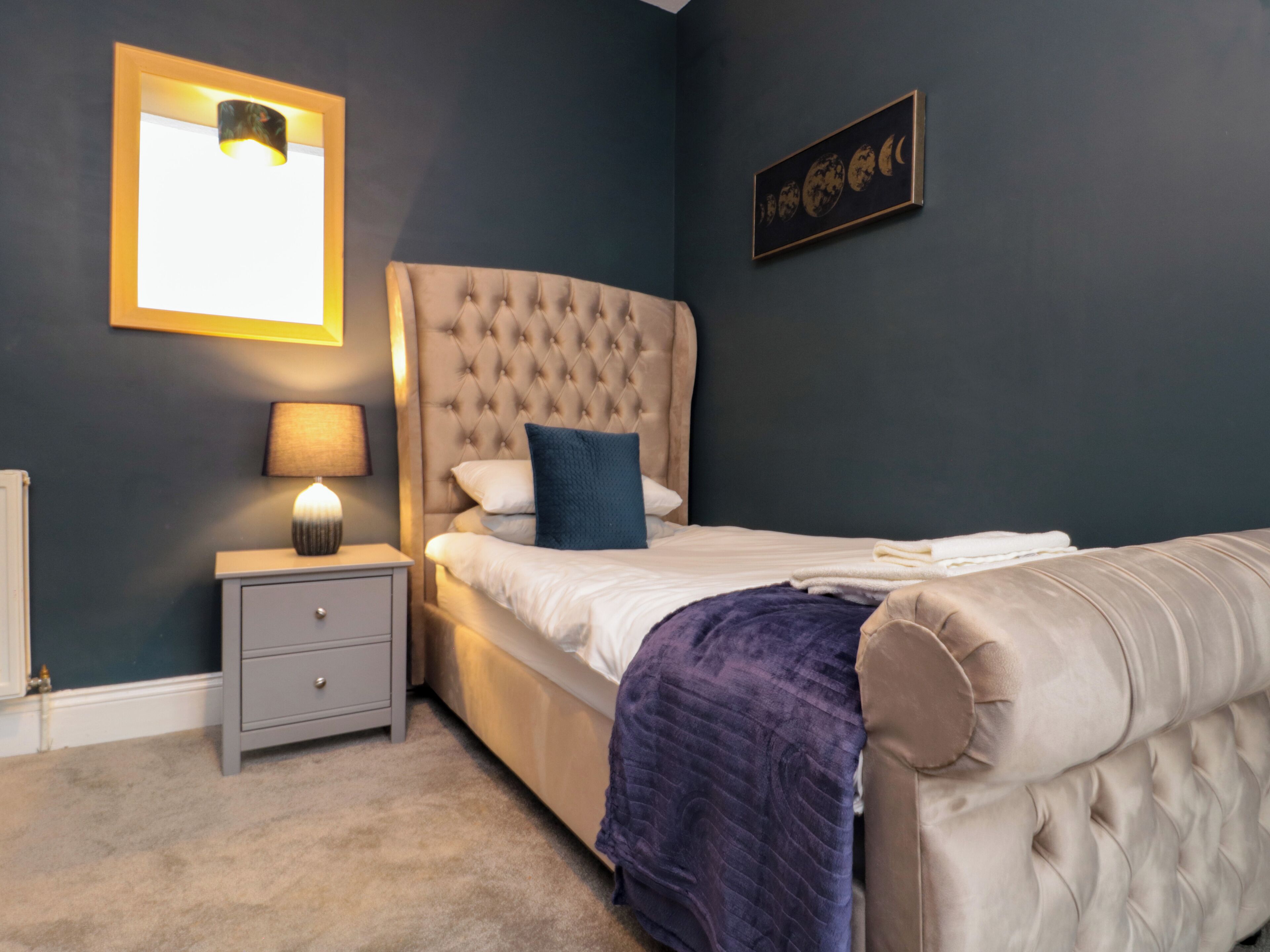 A bedroom with a tufted headboard bed, a bedside table with a lamp, a nightstand, and a wall-mounted picture of the moon phases.