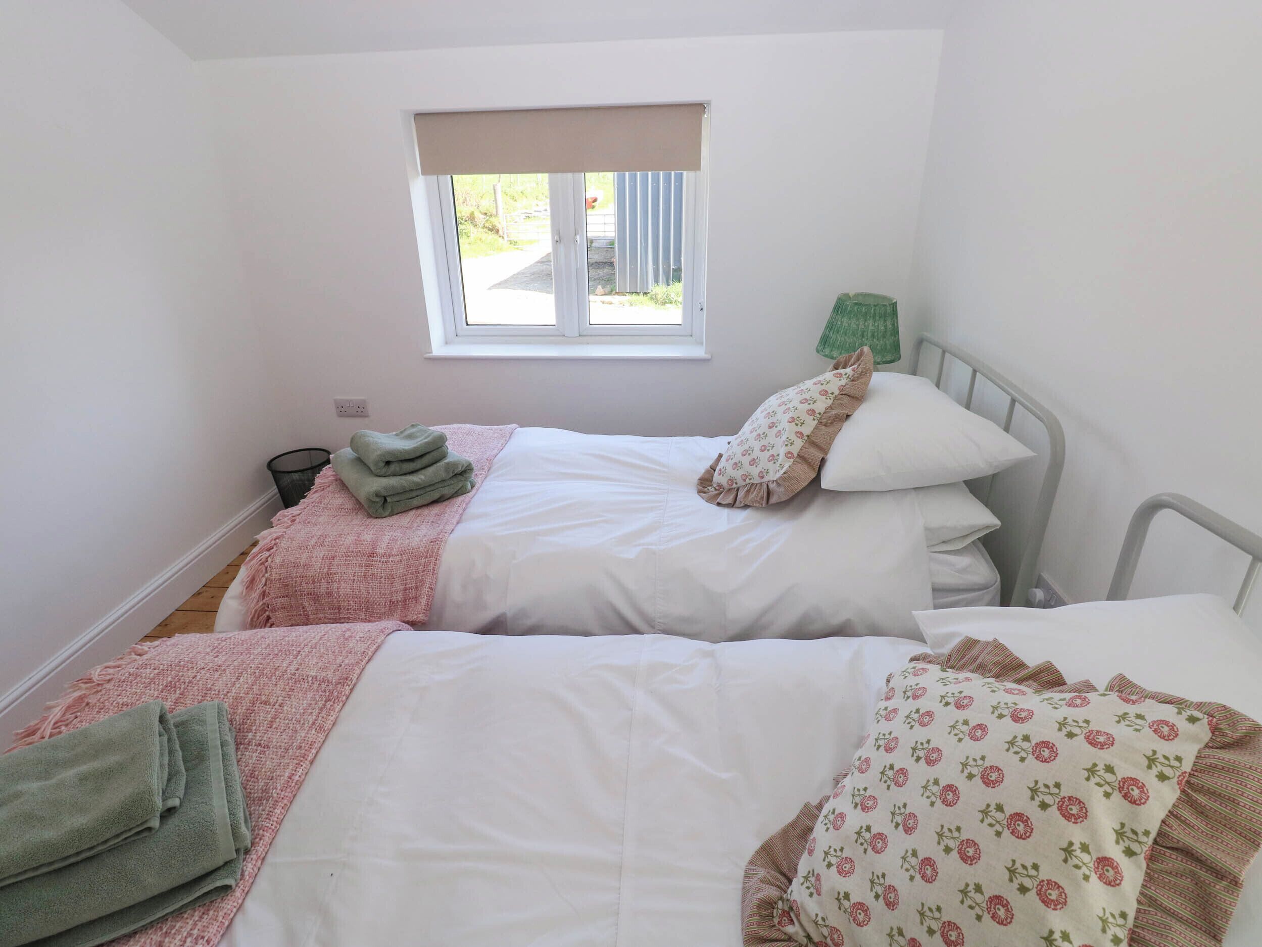 Une chambre avec deux lits, une fenêtre avec un store beige et une lampe verte placée à côté de l’un des lits.