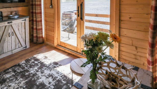 A rustic wooden interior with a kitchen, a dining area with a table and chairs, and a large window with a view of a farm. - Rocky Ridge Cabin (Limavady)