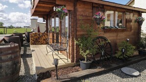 A rustic wooden building with a wooden deck, a metal bench, a large wheel, and flower baskets. - Rocky Ridge Cabin (Limavady)