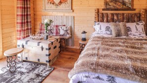A cozy cabin room with a wooden interior, a bed with a fur throw, a chest of drawers, a table with a vase of flowers, and a painting of deer on the wall. - Rocky Ridge Cabin (Limavady)