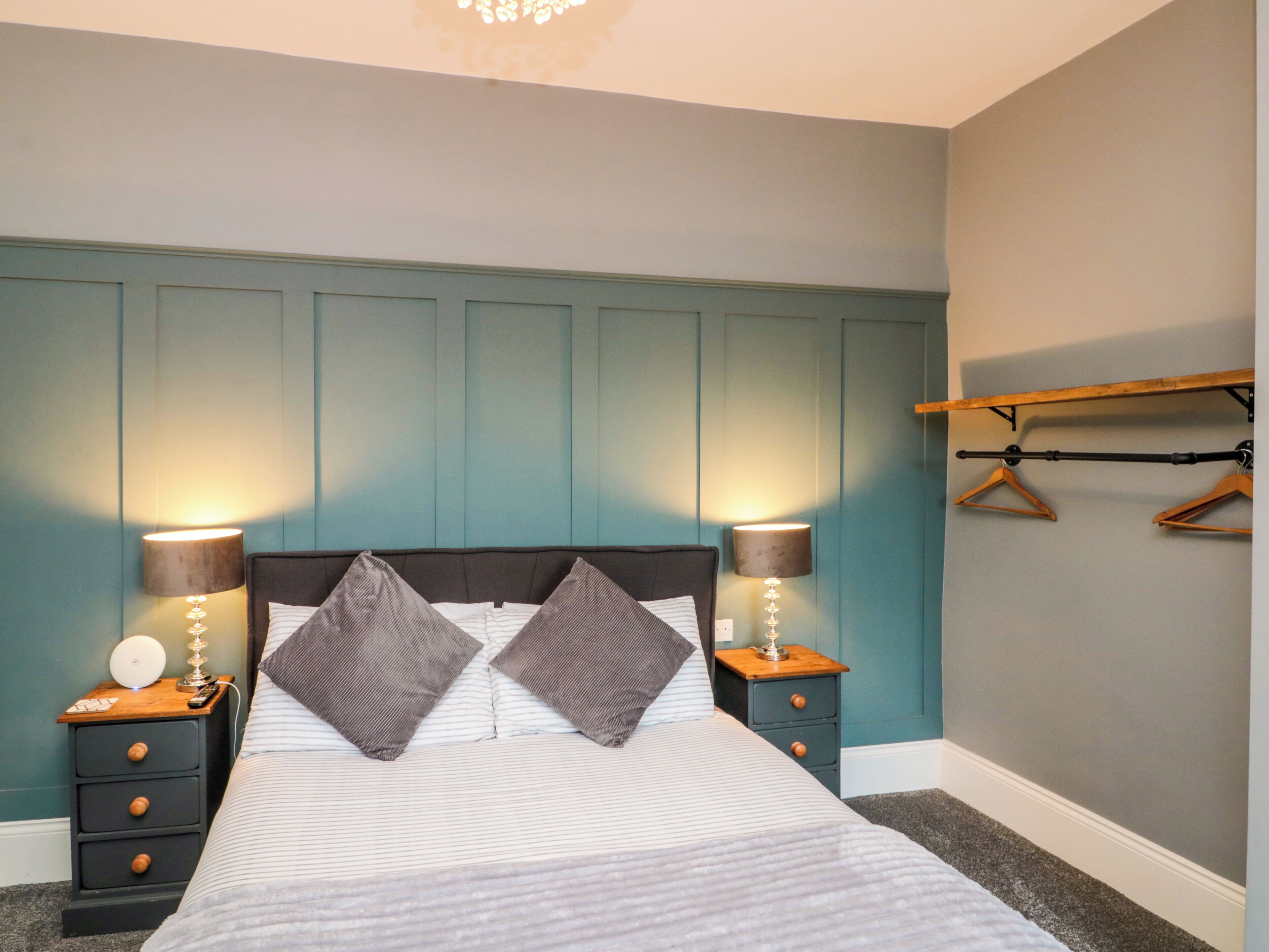 A bedroom with a bed, two bedside tables, a lamp, a hanging clothes rack, and a wall-mounted shelf.