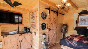 Interior rústico con una mesa de comedor de madera, una cama con manta escocesa, un televisor montado y un reloj en la pared.