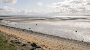 Paisaje costero con playa arenosa, afloramientos rocosos y una amplia extensión de agua bajo un cielo parcialmente nublado.