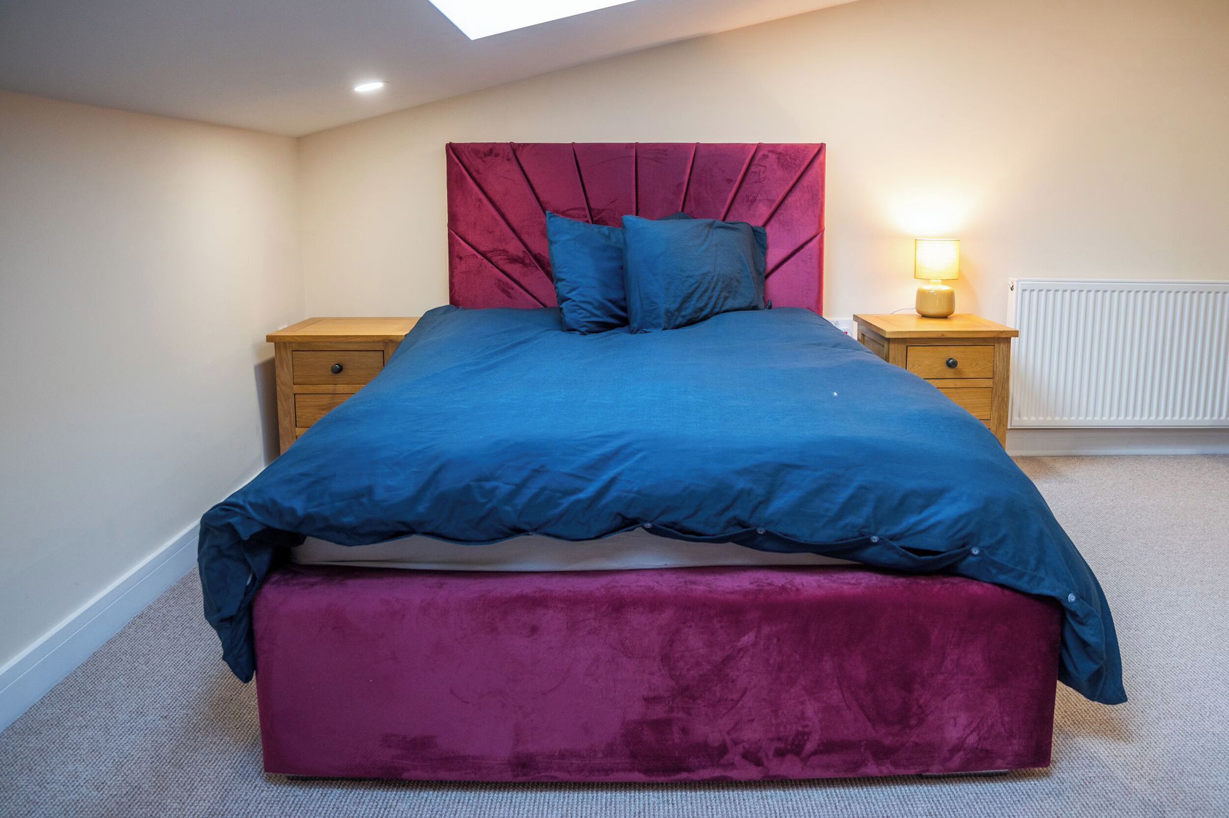 A bedroom with a bed, a bedside table, a lamp, a radiator, and a skylight.