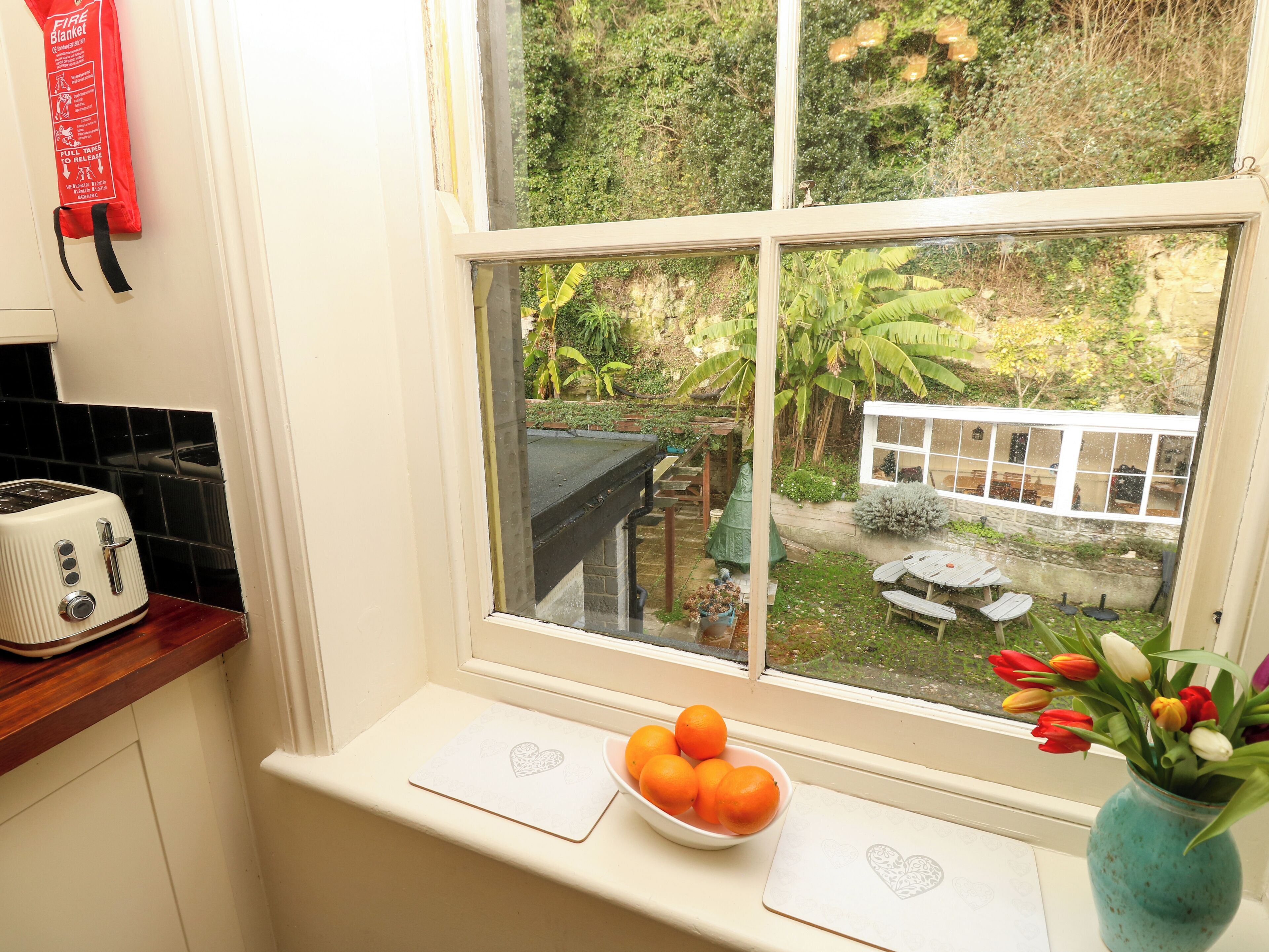 A kitchen with a white countertop, a toaster, a window with a view of a garden, and a vase with flowers on the windowsill.