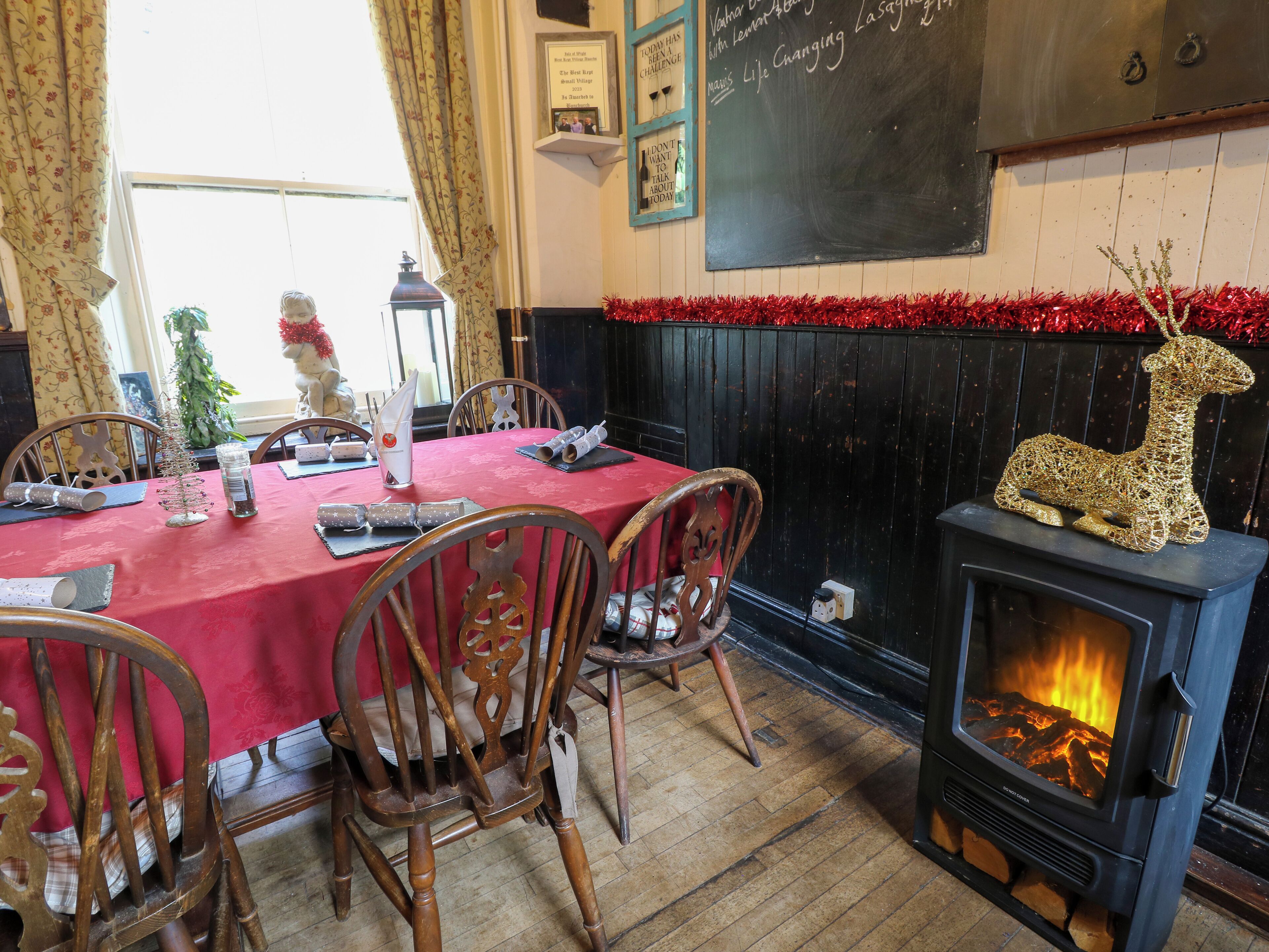 Un comedor acogedor con mantel rojo, sillas de madera, chimenea con un reno decorativo y una pizarra con escritura en la pared.