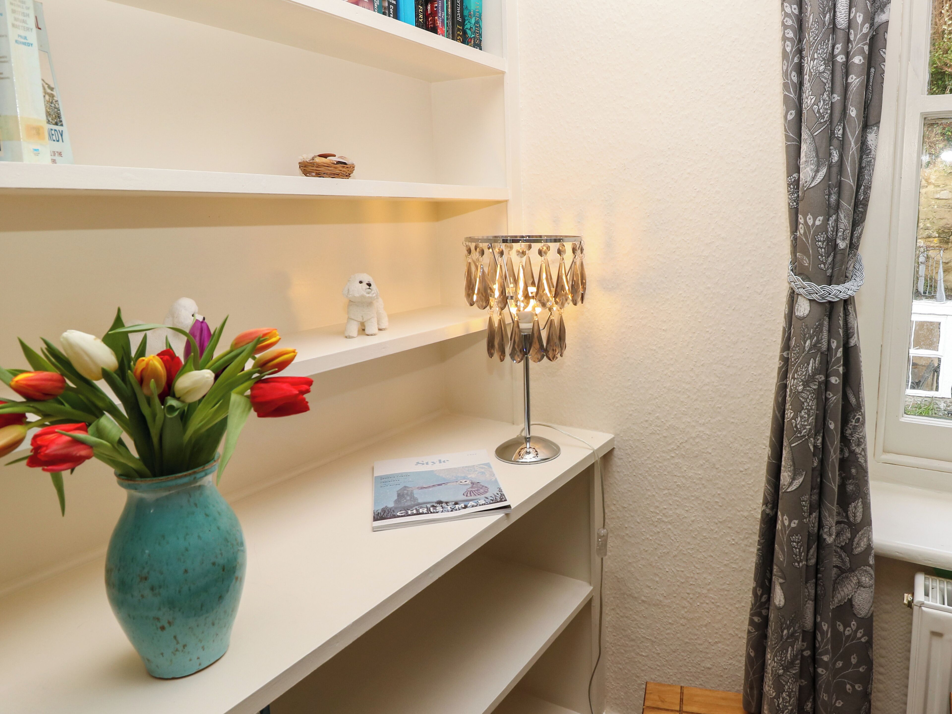 A room with a white shelf holding books, a vase with tulips, a small dog figurine, and a table lamp.