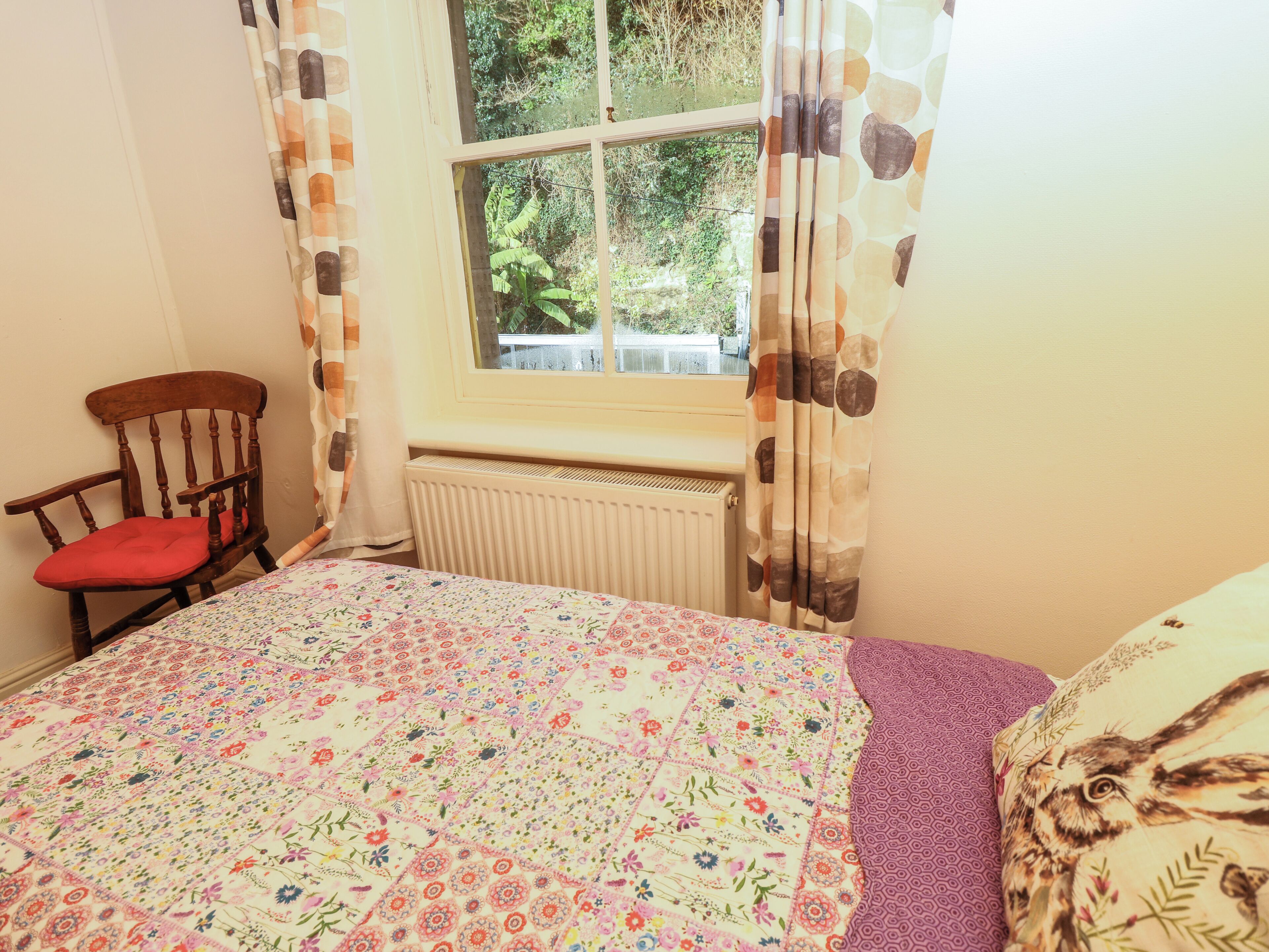 A bedroom with a floral quilt, a wooden chair, a window with curtains, and a radiator.