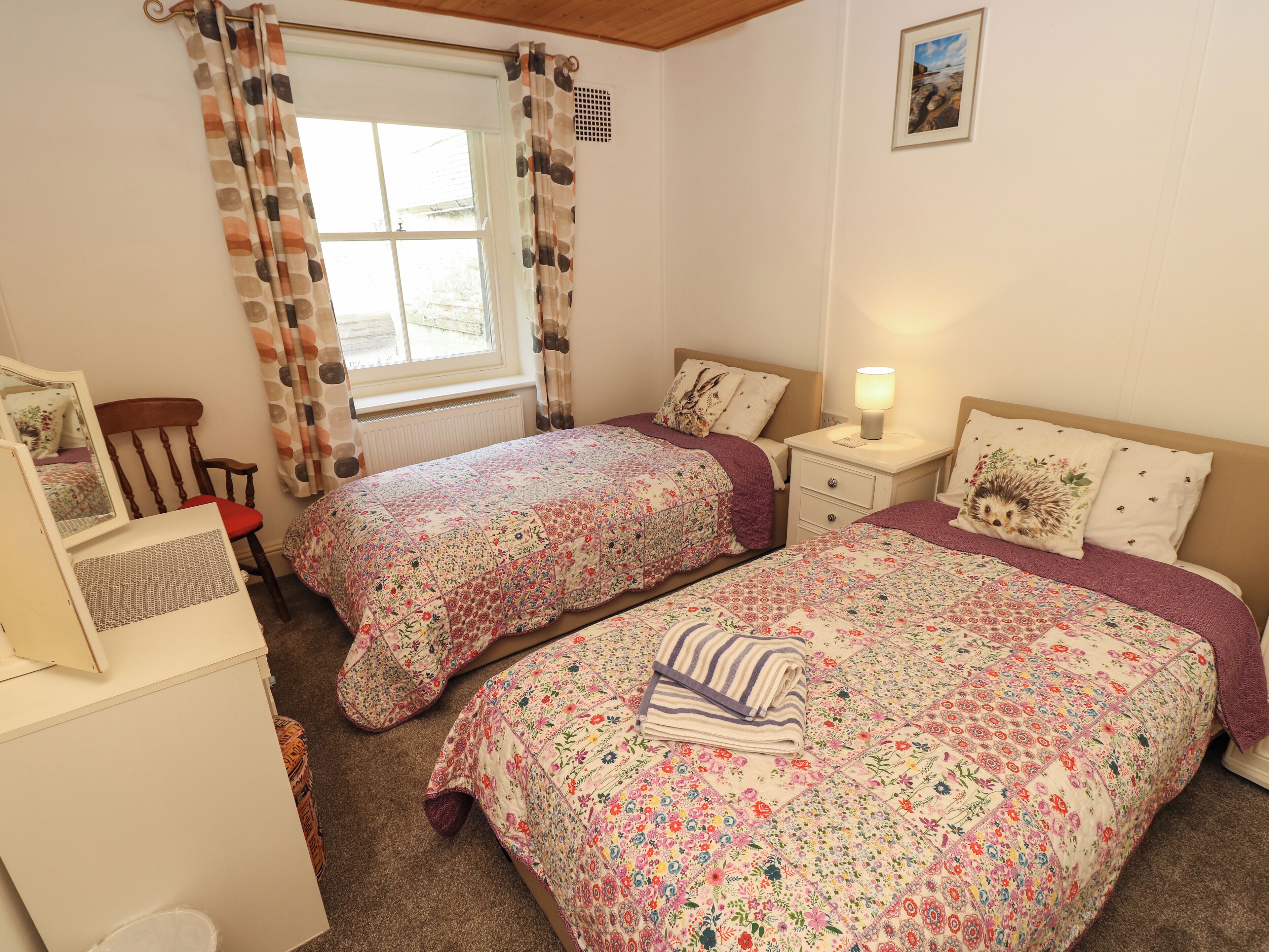 A bedroom with two beds, a dresser, a chair, a window with curtains, a bedside table with a lamp, and a framed picture on the wall.