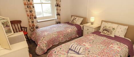 A bedroom with two beds, a dresser, a chair, a window with curtains, a bedside table with a lamp, and a framed picture on the wall.