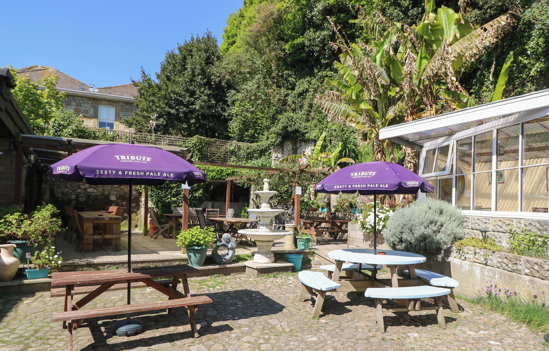 Een terras buiten met picknicktafels en paarse parasols, omgeven door groen en een stenen muur.