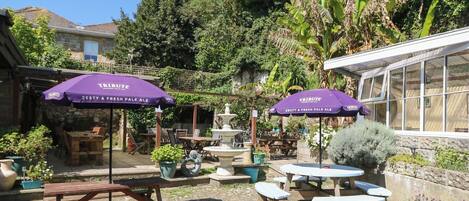 Een terras buiten met picknicktafels en paarse parasols, omgeven door groen en een stenen muur.