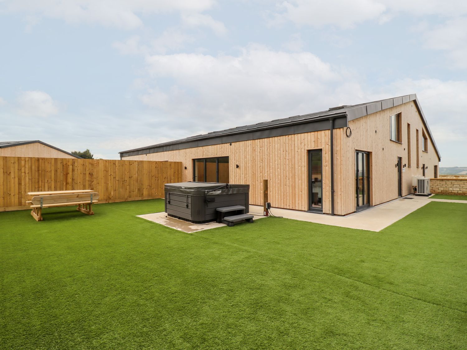 Un edificio moderno con rivestimento in legno, una vasca idromassaggio e un tavolo da picnic in un giardino erboso.