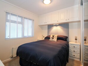 A bedroom with a large bed, white cabinets, a radiator, and a window with blinds. - Un Diwrnod The Warren T22 (Pwllheli)