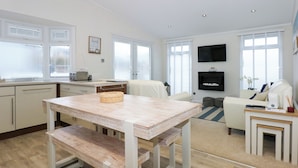 A modern kitchen with white cabinets, a wooden dining table, and a built-in bench. There is a TV mounted on the wall and a window with blinds. - Un Diwrnod The Warren T22 (Pwllheli)