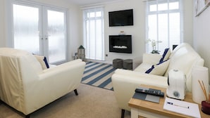 A modern living room with white sofas, a flat-screen TV, and a coffee table with remote controls and a pen. - Un Diwrnod The Warren T22 (Pwllheli)