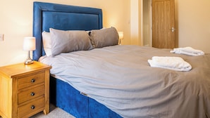A neatly made bed with a blue headboard, a wooden nightstand with drawers, and a lamp. - Bears Court Barn 6 (Cheltenham)